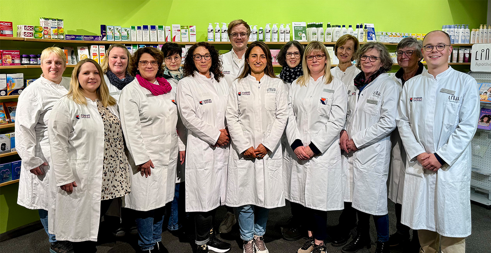 Gruppenfoto eines Apothekenteams mit 14 Personen, die alle weiße Kittel tragen. Sie stehen in einer Reihe vor Regalen mit Pflege- und Gesundheitsprodukten. Alle blicken in die Kamera und lächeln.